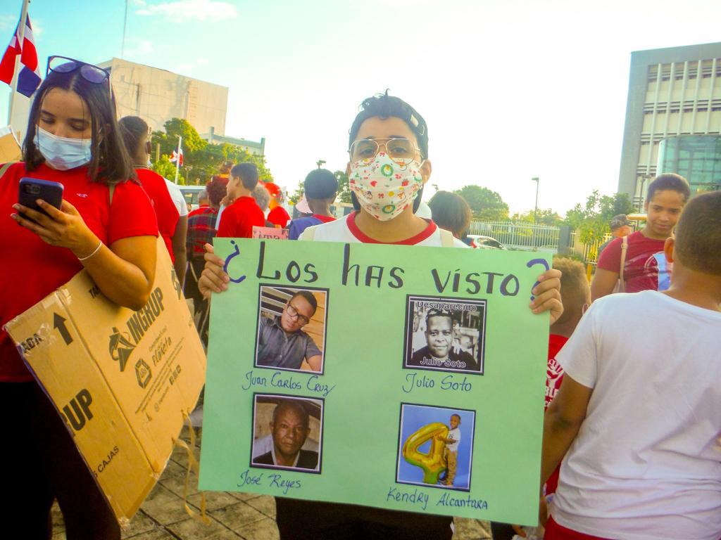 A picture of a masked person holding up a sign that says have you seen them? The sign includes photos of four people, named Juan Carlos Cruz, Julio Soto, Jose Reyes, and Kendry Alcantara